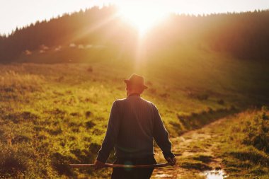 Gün batımında kırsal bir yolda yürüyen biri elinde sopayla arka planda orman tepesi olan bir adam..