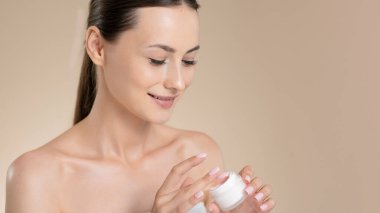 Mutlu genç bir kadın bej rengi stüdyo geçmişine krem sürüyor. Çıplak omuzlu güzel esmer nemlendirici cilt için kozmetik kullanıyor. Güzellik prosedürleri ve kaplıca konsepti.