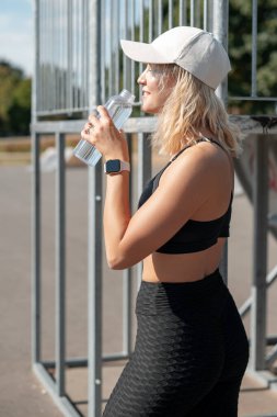 Sarışın kadın güneşli bir günde antrenmandan sonra dinlenirken stadyumda su içiyor. Sokak çalışması ve su dengesi kavramı