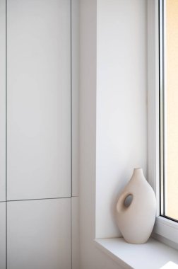Modern ev dekorasyonu, ürün sahneleme veya yaşam tarzı kavramları için ideal, minimalist bir iç mekanda, temiz beyaz bir pencere eşiğine yerleştirilmiş zarif, nötr seramik vazo..