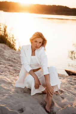 Göl kenarında kumların üzerinde oturan şık beyaz takım elbiseli etkili genç sarışın. İnanılmaz günbatımının bulanık arkaplanı. Moda ve güzellik kavramı.