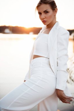 Kendine güvenen, modaya uygun beyaz takım elbiseli genç bir kadın arkasında muhteşem bir günbatımı olan kameraya bakıyor. Doğal güzellik ve moda anlayışı.