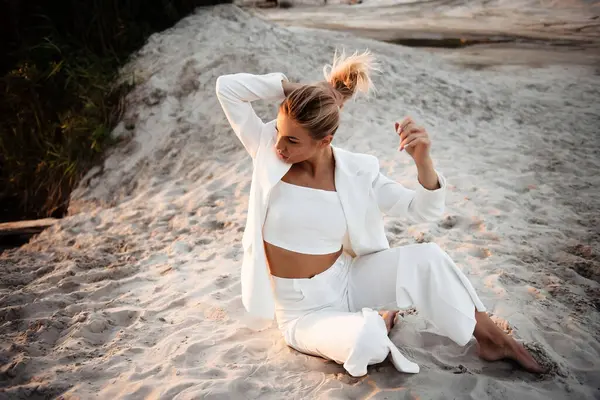 Güzel, beyaz takım elbiseli bir bayan kumların üzerinde oturuyor, sarı saçlara dokunuyor ve yan bakıyor. Mutlu kadın gün batımında doğada fotoğraf çekimi yapıyor..