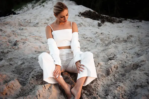 Tasarımcı beyaz takım elbiseli profesyonel model yaz sezonunda dışarıda çalışıyor. Sarı saçlı genç bir kadın çıplak ayakla sıcak kuma dokunuyor..