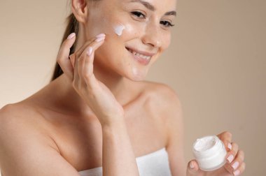 Stüdyoda yüzüne krem süren çıplak omuzlu şehvetli genç bir kadın. Beyaz kadın model bej arka planda güzellik ayinlerinin tadını çıkarıyor. Taze doğal ten..