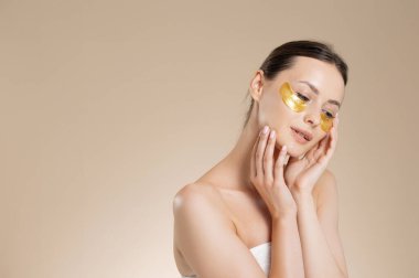 Çıplak omuzlu narin genç bir kadın gözlerinin altında altın bantlar kullanıyor. Bej renkli stüdyo arka planında izole edilmiş. Güzellik ve cilt bakımı.