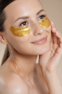 Gözlerinin altında kolajen altın pedlerle bej rengi stüdyo arkasında poz veren pozitif genç bayan. Güzel model güzellik prosedürlerini seviyor. Cilt bakımı kavramı.