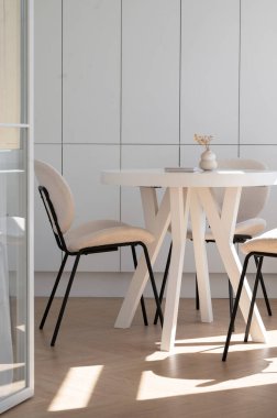Minimalist yemek alanı, beyaz yuvarlak masa, bej sandalyeler ve masada küçük bir vazo. Güneş ışığı, ahşap zemine gölge düşürür. Sıcak ve davetkar bir atmosfer yaratır..