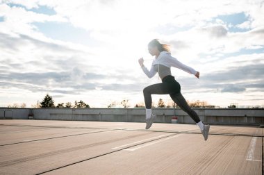 Atletik kıyafetli bir kadın, arka planda bulutlu bir gökyüzü ile çatıda koşarken havada yakalanır..