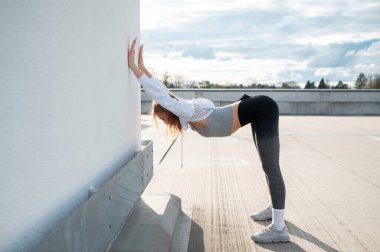 Çatıdaki bir duvara yaslanan genç bir kadın. Uygun bir kıyafet giyiyor, aktif bir yaşam tarzı sergiliyor. Gökyüzü parçalı bulutlu, parlak bir atmosfer yaratıyor..