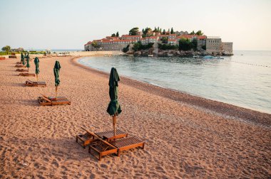 Boş plaj, ahşap şezlonglar ve tarihi bir ada kasabasına bakan kapalı şemsiyeler..
