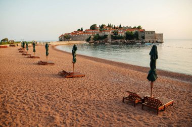 Tarihi bir ada oteline bakan güneşli şezlongları ve şemsiyeleri olan boş bir sahil..
