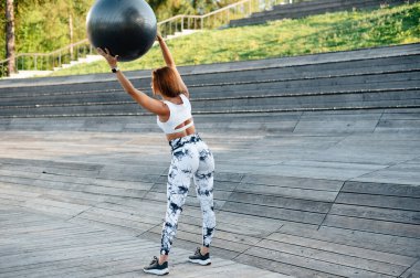 Atletik kıyafetli bir kadın dışarıda egzersiz yapıyor, kafasının üstüne büyük bir egzersiz topu kaldırıyor. Bir parkta tahta basamaklarda, dayanıklılık ve zindelik gösteriyor..