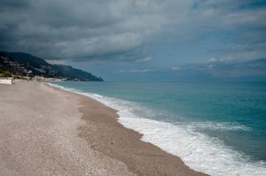 Sakin bir sahil, bulutlu bir gökyüzünün altında hafif dalgalar, arka planda dağlar..