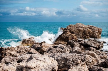 Dalgalar parçalı bulutlu bir gökyüzünün altında kayalık bir sahil şeridinde büyük kayalara çarpıyor..