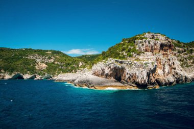 Berrak mavi gökyüzünün altında kayalık kayalıkları ve yemyeşil yeşillikleri olan manzaralı bir kıyı manzarası..