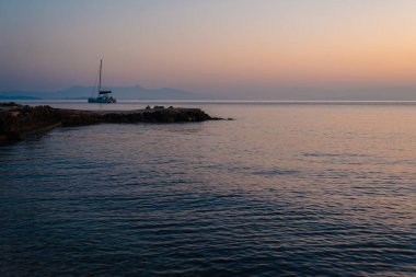 Sakin bir denizin üzerinde sakin bir günbatımı, uzakta bir yelkenli, huzurlu ve sakin bir atmosfer yaratıyor..