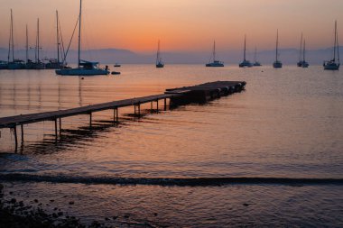 Sakin bir denizin üzerinde sakin bir günbatımı. Tahta bir iskelenin yanında demirli yelkenliler..