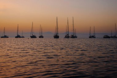 Gün batımında sisli gökyüzü ile sakin sularda yüzen siluetli yelkenliler..