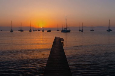 Sakin bir denizin üzerinde sakin bir gün batımı siluetli yelkenliler ve ahşap iskele.