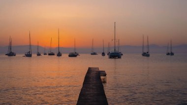 Sakin bir denizin üzerinde sakin bir gün batımı. Tahta bir iskelenin yanında demirli yelkenliler..