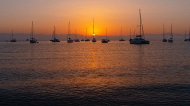 Sakin bir denizin üzerinde sakin bir gün batımı ufukta siluetli yelkenliler.
