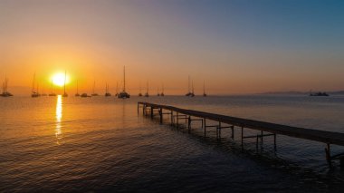 Sakin bir denizin üzerinde sakin bir günbatımı. Suya uzanan ahşap bir iskele ve ufukta süzülen yelkenliler..