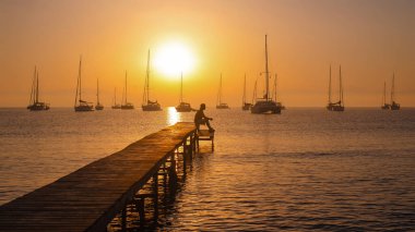 Sakin bir denizin üzerinde sakin bir günbatımı ve uzak bir yere demir atmış yelkenliler. Bir insan, altın gökyüzüne karşı silueti olan ahşap bir iskelenin ucunda oturuyor..