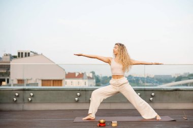 Sarışın kadın sabahları çatıda yoga pozu veriyor. Beyaz kıyafetli kıvırcık kadın, arka planda yaz manzarası olan modern binanın çatısında kollarını spor minderine açmış Asana 'yı canlandırıyor.