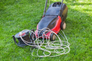 Yeşil bir çimenlikteki kırmızı elektrikli çim biçme makinesi. Kablolar, elektrik uzatma kablosu, taşınabilir soket.