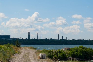 Fotoğraf endüstriyel bir manzarayı gösteriyor. Ön planda vahşi doğa, arka planda ise bir endüstriyel kompleks. Gökyüzü beyaz bulutlarla mavidir.