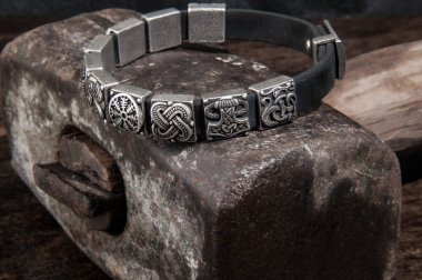 : Üzerinde Viking sembolleri olan gümüş bir bileziğin yakın çekimi, ağır metal bir çekiç üzerinde duruyor..