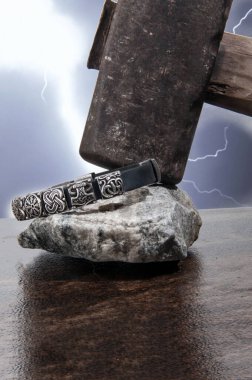 Üzerinde Viking sembolleri olan gümüş bir bileziğin yakın çekimi fırtınalı bir gökyüzüne karşı Thor 'un çekicinin üzerinde duruyor..