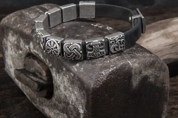 : Üzerinde Viking sembolleri olan gümüş bir bileziğin yakın çekimi, ağır metal bir çekiç üzerinde duruyor..