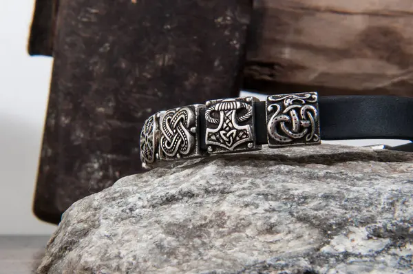 Üzerinde Viking sembolleri olan gümüş bir bileziğin yakın çekimi fırtınalı bir gökyüzüne karşı Thor 'un çekicinin üzerinde duruyor..