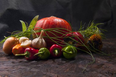 Kırmızı balkabağı, canlı biberler, sarımsak kafalar, soğanlar ve taze yeşilliklerle dolu zengin bir sonbahar hayatı. Hepsi koyu desenli ahşap bir yüzeye yerleştirilmiş..