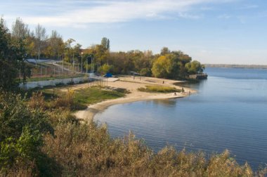 Kumlu ve sakin suyu olan bir sonbahar kumsalı manzarası. Kıyı boyunca ağaçlar sarı yapraklarla kaplıdır. Şehir manzarasının üzerindeki açık gökyüzü.