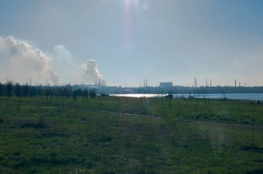 Ufukta dumanı tüten bir endüstriyel kompleksi olan geniş bir alan ve su manzarası. Doğanın ve şehirleşmenin etkileşimini sembolize eder, metin için alan yaratır.