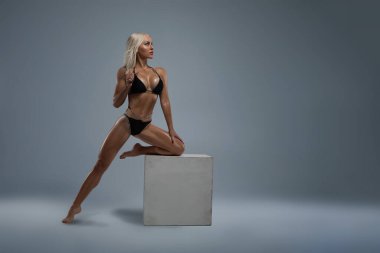 Stüdyoda beyaz bir arka planda siyah mayo giymiş güzel, bronzlaşmış sarışın kız fitness modeli dizini bir küp üzerinde incelikle dinlendiriyor ve seksi bir kadın vücudunu gösteriyor.