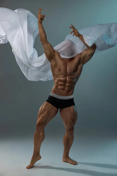 Aesthetics of the male athletic body. The male model pulls his hands up, standing on his toes. White fabric covers male muscular body
