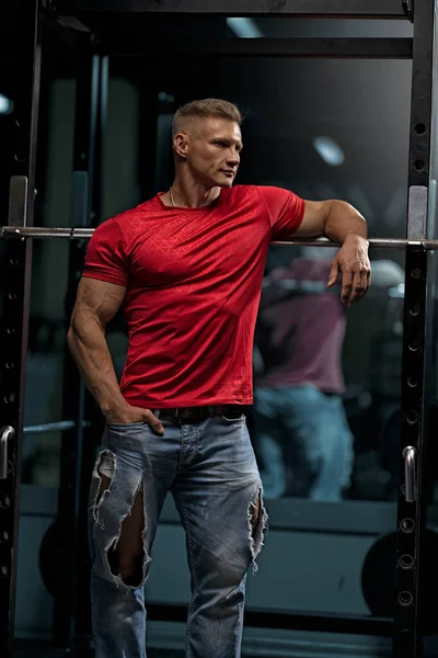 A well-built athletic young adult male in a red t-shirt and ripped jeans poses in the gym next to the barbell at Smiths barre. Fitness trainer with big muscles