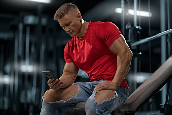 Strong athletic male trainer in jeans and a red t-shirt sits on a bench in the gym and looks at the phone screen