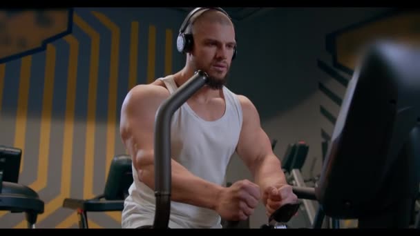 一个戴着耳机 体形匀称的秃头肌肉男和一个白色油箱盖在体育馆的台阶上做有氧运动 在有氧运动过程中展示健美运动员特写的形象 — 图库视频影像