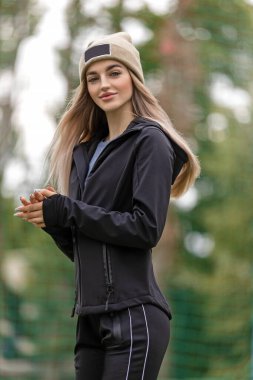 Büyük dudaklı ve mavi gözlü genç bir kadın çerçeveye bakar, siyah bir spor ceketin kelepçelerine dokunur. Sokaktaki şapkalı güzel sarışın kadın. Spor kıyafetleri.