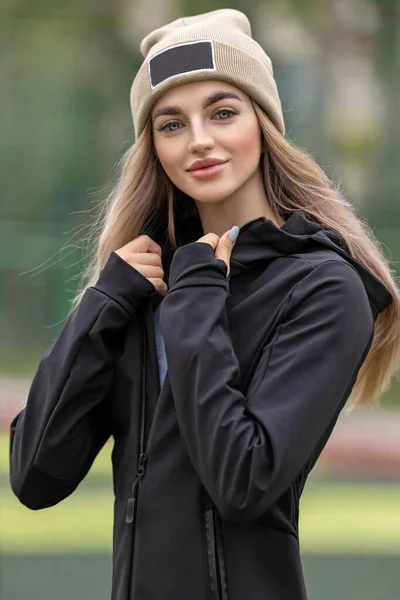 A young woman with big lips and blue eyes looks into the frame, holding on to the collar of a black sports jacket. Beautiful blonde woman in a hat on the street. Sportswear fashion