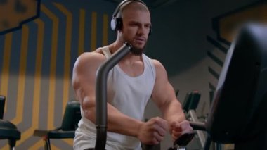 Kulaklıklı, kaslı, kel bir adam ve beyaz kolsuz tişörtlü bir adam spor salonundaki bir basamağa kardiyo yapıyor. Vücut geliştirici figürünün kardiyo sürecinde yakın plan görüntüsü