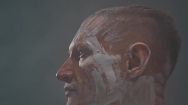 İyi yapılı atletik bir adam duman kullanarak beyaz bir siklonamada poz verir. Yüz ve kaslı erkek vücudu, estetik bir antika heykel gibi beyaz kille kaplıdır..