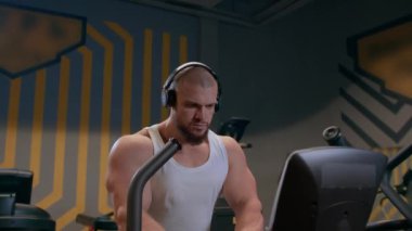 Kulaklıklı, kel, atletik ve beyaz tişörtlü bir adam spor salonunda bir basamağa kardiyo yapıyor. Kuvvet ve motivasyon.