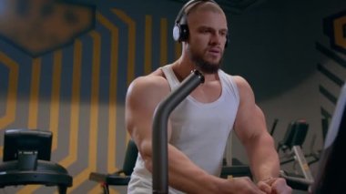 Kulaklıklı, kel, atletik ve beyaz tişörtlü bir adam spor salonunda bir basamağa kardiyo yapıyor. Kuvvet ve motivasyon.