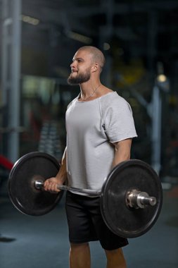 Spor salonunda ağırlık kaldıran kaslı bir adam. Halterli pazılar için dayanıklılık eğitimi. Sakallı kel adam ağır ağır egzersiz yapıyor.